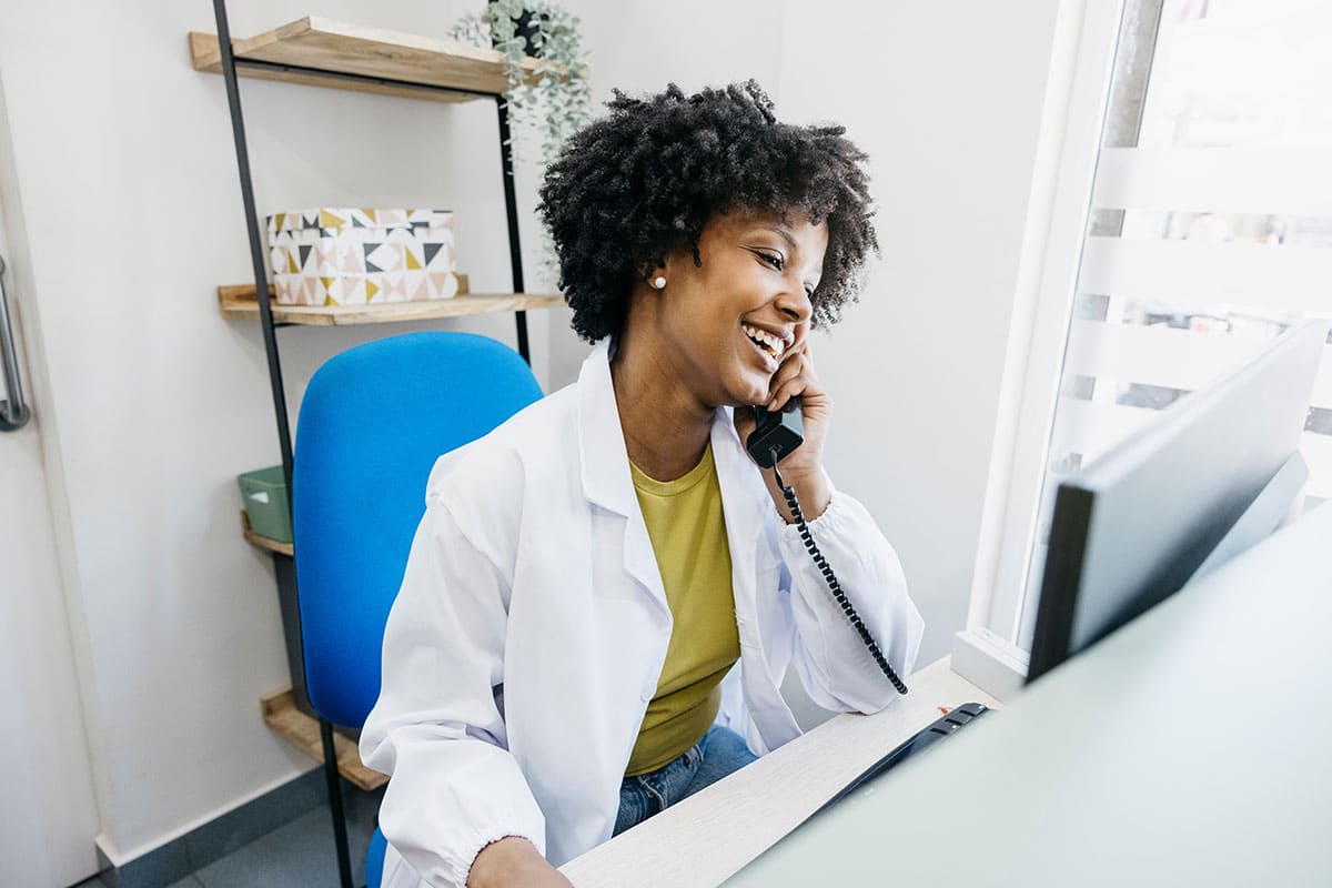 Smiling medical receptionist talking on phone and using computer, managing appointments and providing customer service
